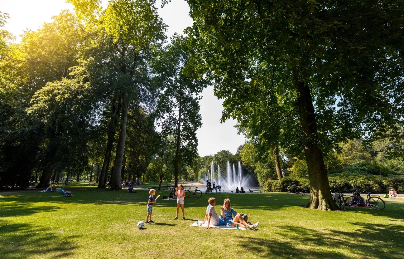 Grass area in Valkenbergpark, surrounded by trees, a family is relaxing in the sun, at the back there is a fountain visible.
