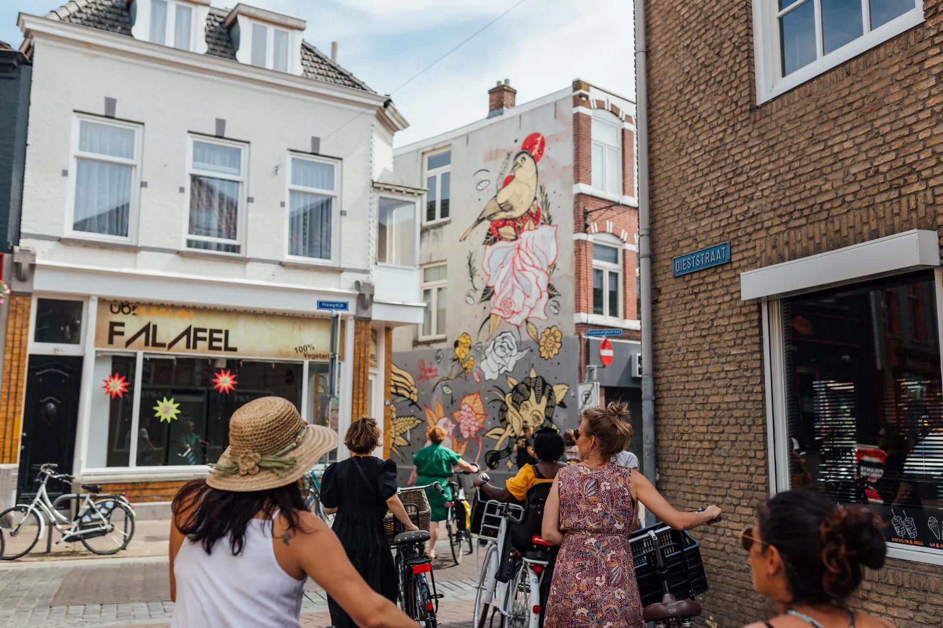 Mensen lopen met de fiets aan de hand door de stad langs een foto van de Blind Walls Gallery.